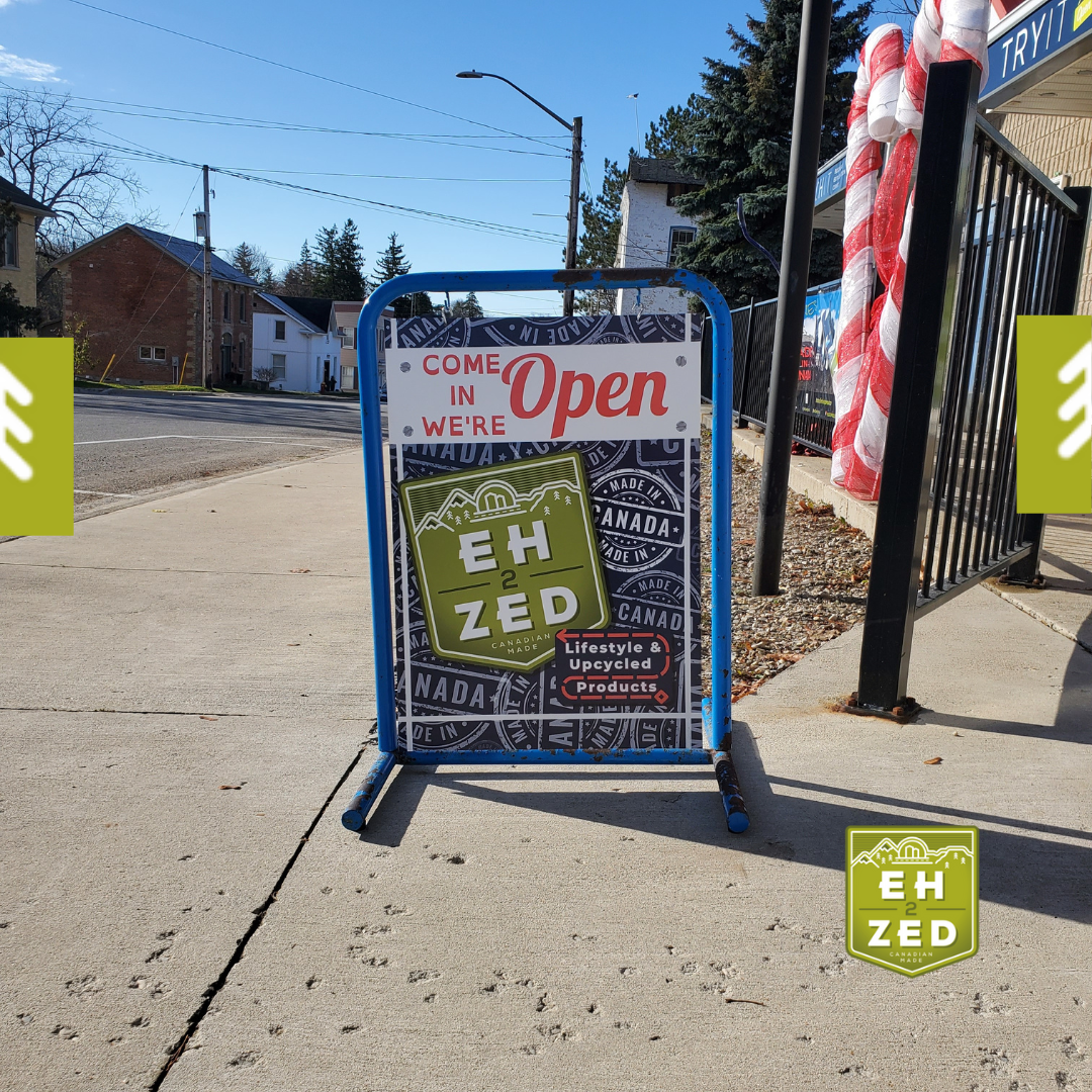 A blue metal open sign for Eh-2-Zed on a sidewalk on a sunny day