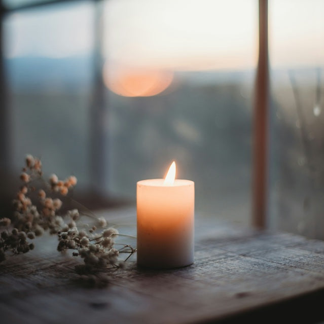 candle on wood surface beside some small flowers infront of a window