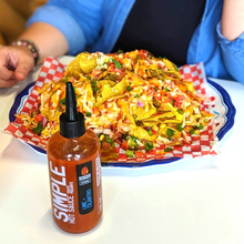 Load image into Gallery viewer, close up of tacos with a bottle labelled hot sauce in front and a person&#39;s chest and hand behind
