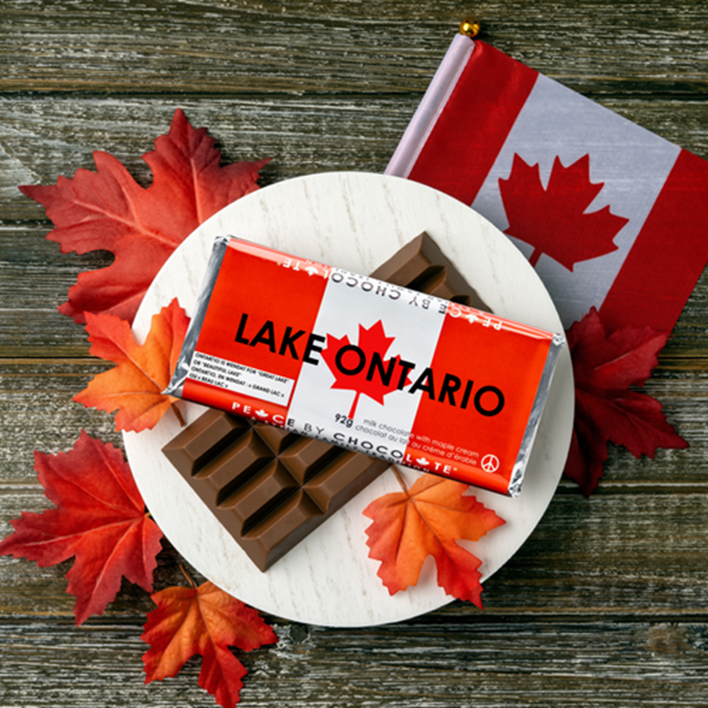 A chocolate bar wrapped in a red and white Lake Ontario Canada flag wrapper, placed on a white plate with autumn maple leaves on a rustic wooden surface. A small Canadian flag is visible in the background.