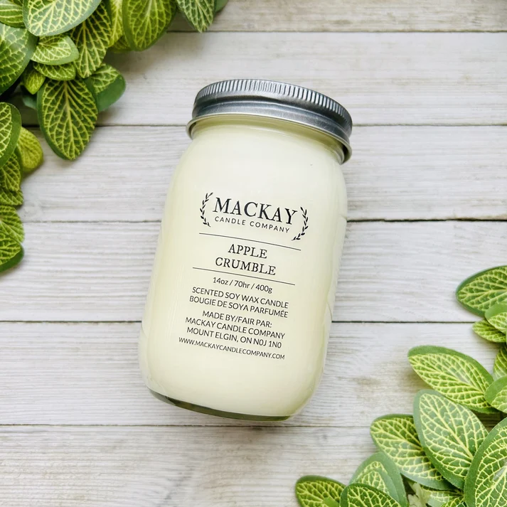 A jar candle labeled 'MACKAY CANDLE COMPANY' and 'APPLE CRUMBLE' sits on a whitewashed wood surface, surrounded by green leafy stems. The candle wax is a pale creamy yellow, and the jar has a silver metal lid.