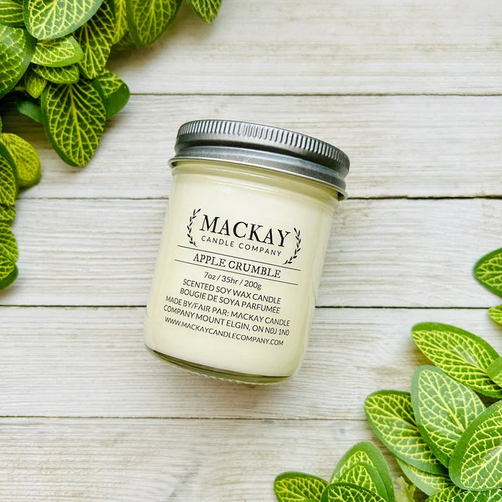 A jar candle labeled 'Mackay Candle Company' and 'Apple Crumble' sits on a whitewashed wood surface, surrounded by green variegated leaves. The candle has a cream-colored wax and a silver metal lid.