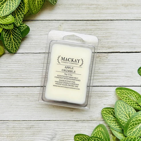A packaged wax melt from Mackay Candle Company labeled 'Apple Crumble' sits on a whitewashed wood surface with green plant leaves around it.