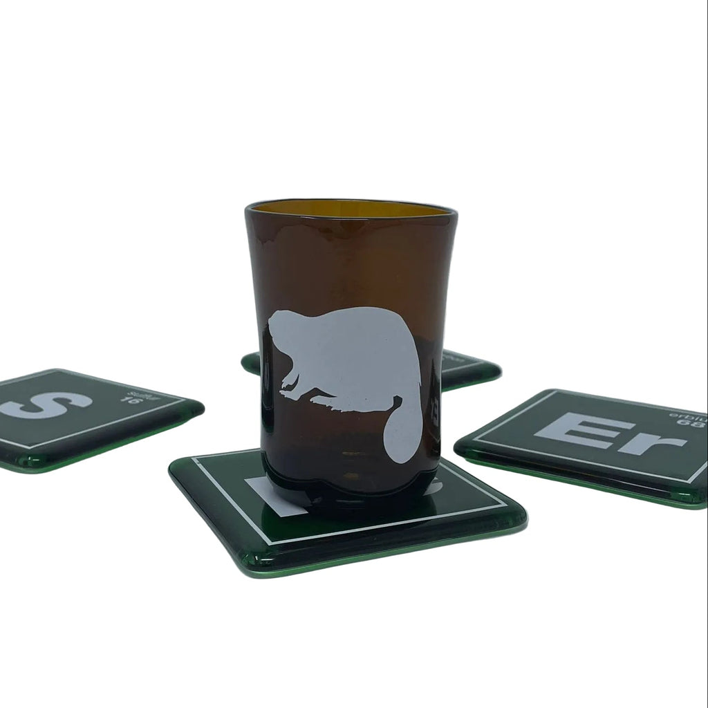 A recycled beer bottle amber brown glass with a white beaver silhouette, resting on a dark green square coaster. Four matching dark green coasters are arranged on a white surface against a light gray background.