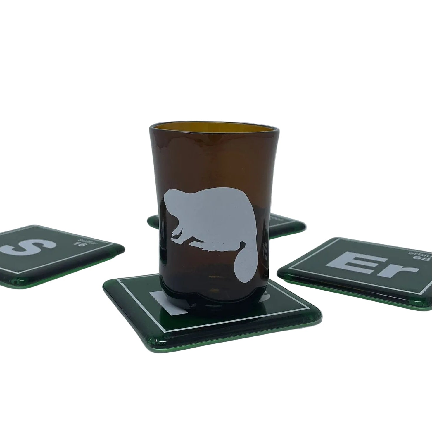 A recycled beer bottle amber brown glass with a white beaver silhouette, resting on a dark green square coaster. Four matching dark green coasters are arranged on a white surface against a light gray background.