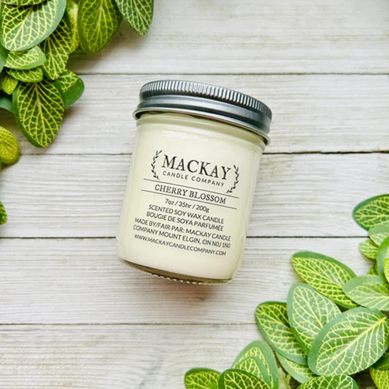 Jar of Mackay Candle Company cherry blossom candle on a wooden surface with green leaves.