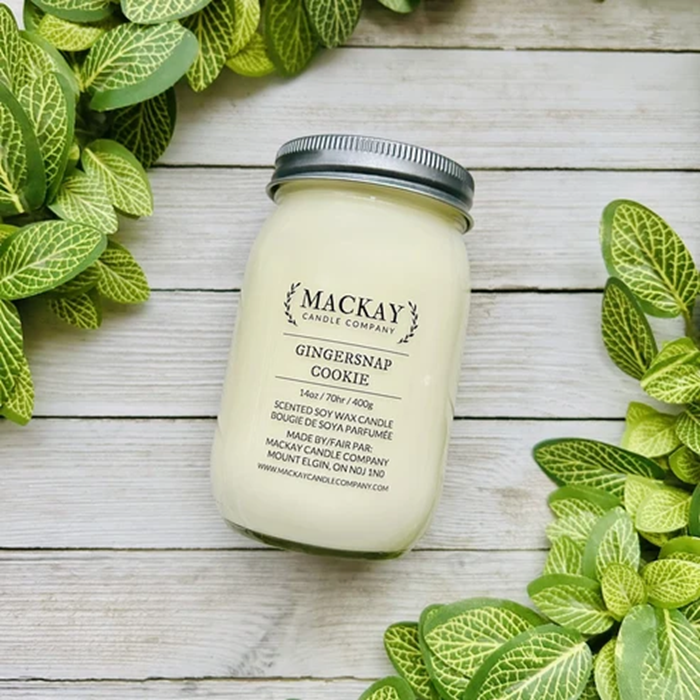 A jar candle labeled 'MACKAY Candle Company Gingersnap Cookie' sits on a whitewashed wood surface surrounded by green leaves. The candle wax is a pale creamy yellow and the jar has a silver metal lid.