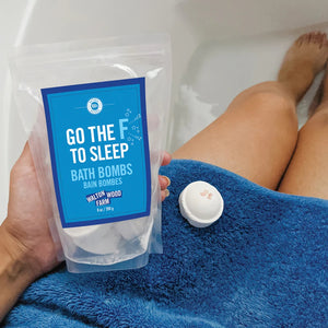 person holding clear plastic bag with blue label filled with bath bombs while sitting on edge of tub with blue towel