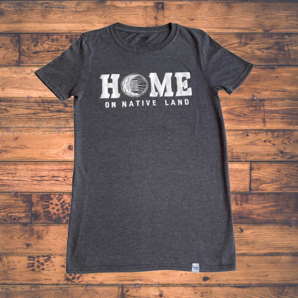 Dark gray t-shirt with 'HOME ON NATIVE LAND' text on a wooden surface