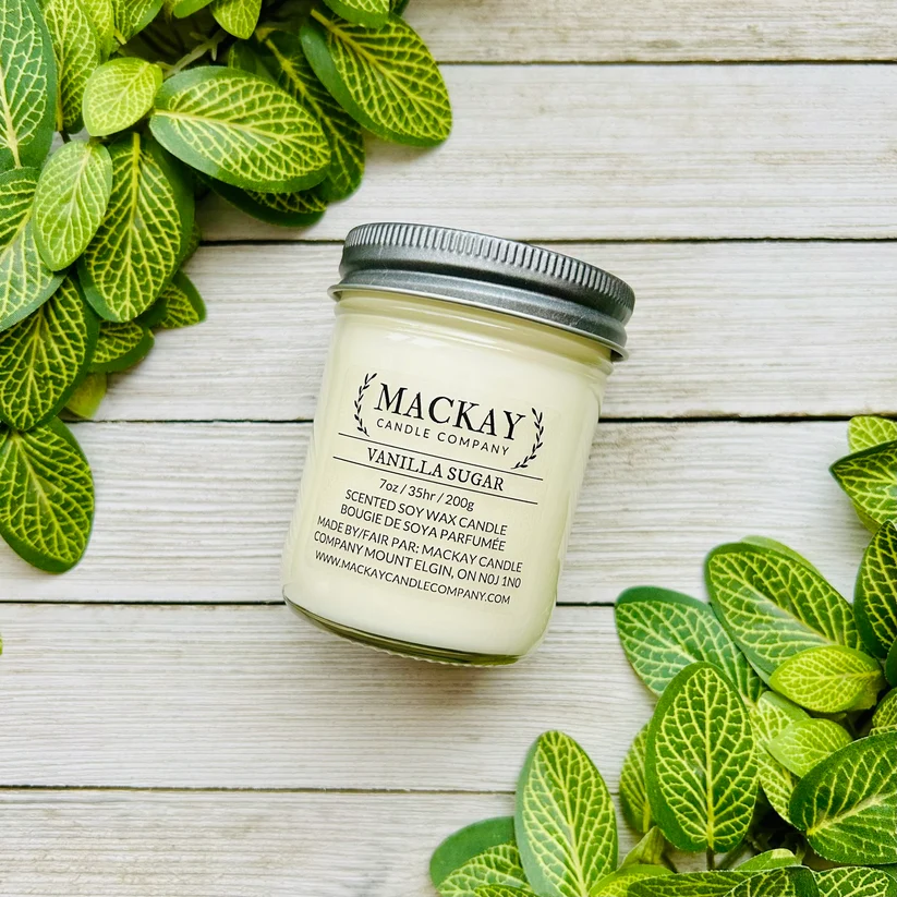 A jar candle labeled 'MACKAY Candle Company Vanilla Sugar' sits on a whitewashed wood surface surrounded by green variegated leaves. The candle jar has a silver metal lid and cream-colored wax.