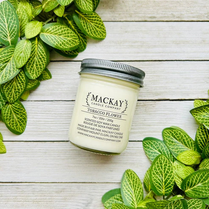 Jar of 'Tobacco Flower' candle from Mackay Candle Company on a wooden surface with green leaves.