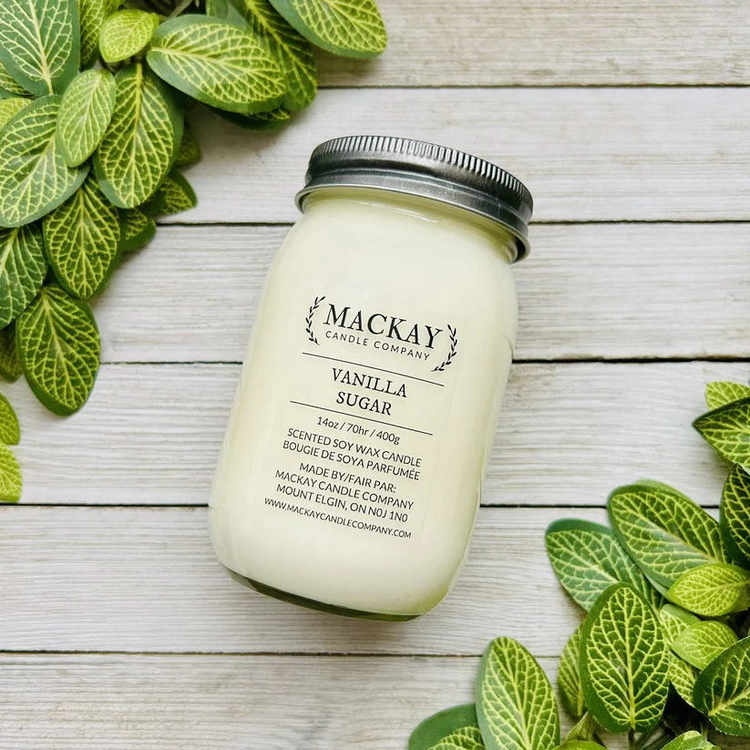 A jar candle labeled 'MACKAY CANDLE COMPANY Vanilla Sugar' sits on a whitewashed wood surface surrounded by green variegated leaves. The candle is in a clear glass jar with pale creamy wax and a silver metal lid.