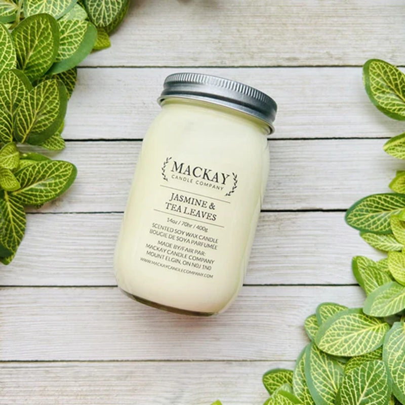 Mackay Candle Company candle with jasmine and tea leaves scent on a wooden surface with green leaves.