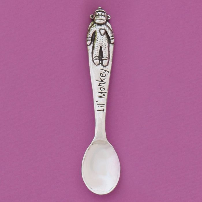 A silver spoon with a monkey figure at the end of the handle and the text 'Lil' Monkey' engraved on the handle, set against a solid pink background.