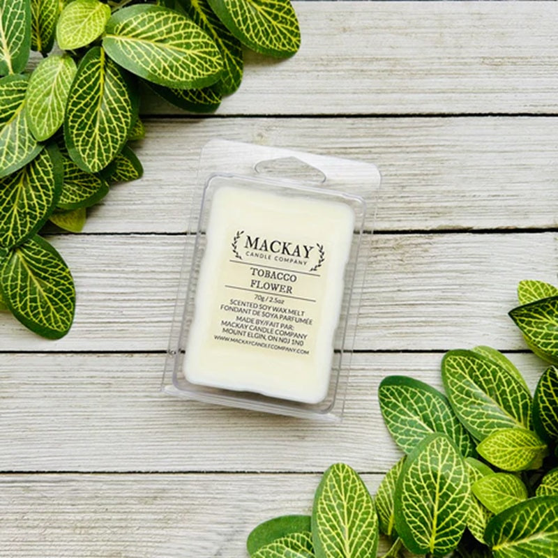 Mackay Candle & Soap Tobacco Flower candle in packaging on a wooden surface with green leaves.