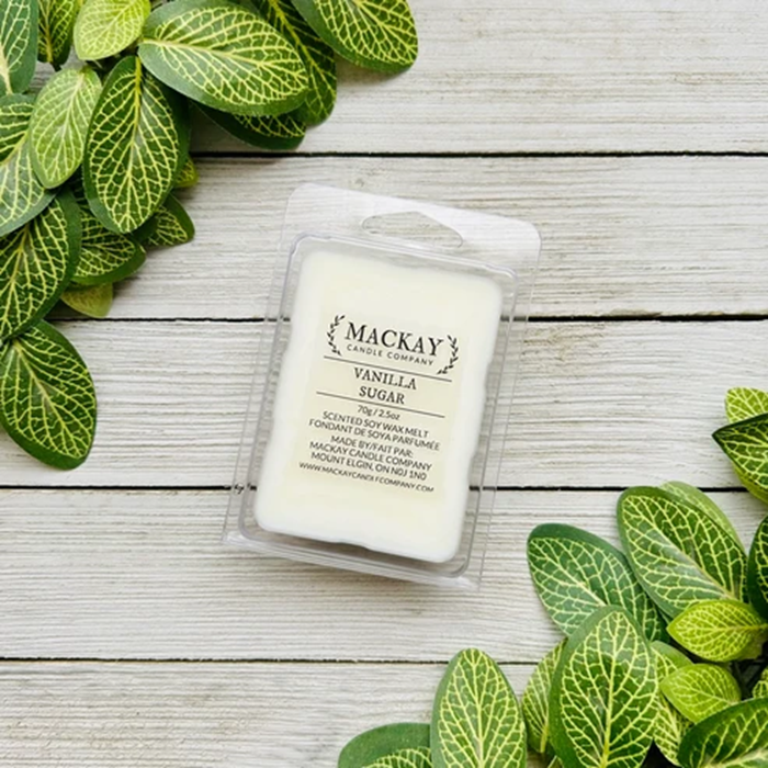 A packaged wax melt labeled 'MACKAY SCENTS COMPANY VANILLA SUGAR' sits on a light wood surface, surrounded by green trailing plant leaves. The product is shown in a clear plastic clamshell with a white wax melt visible inside.