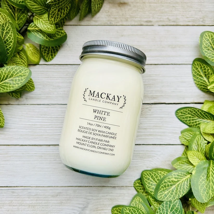A white soy wax candle in a clear glass mason jar with a silver metal lid, labeled 'MACKAY CANDLE COMPANY' and 'WHITE PINE'. The jar is styled on a white wood plank surface, framed by green and white variegated leaves.