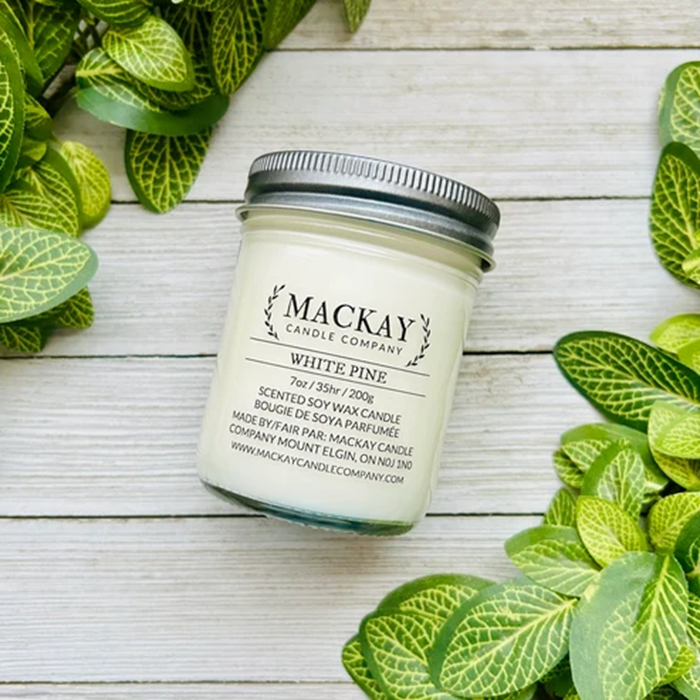 A jar candle labeled 'MACKAY CANDLE COMPANY' and 'WHITE PINE' sits on a whitewashed wood surface surrounded by green variegated leaves. The candle has a white wax fill, a clear glass jar, and a silver metal lid.