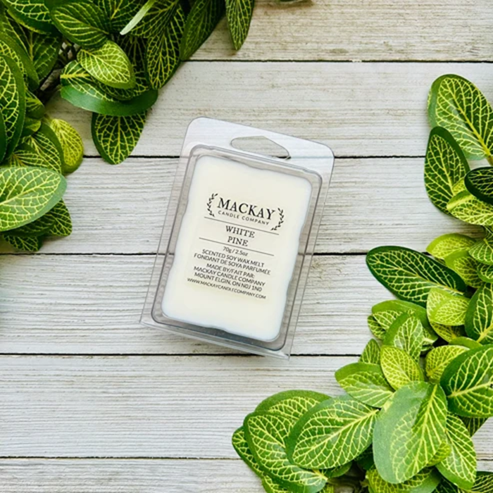 A wax melt package labeled 'MACKAY Candle Company' and 'WHITE PINK' sits on a whitewashed wood surface, surrounded by green leaves.