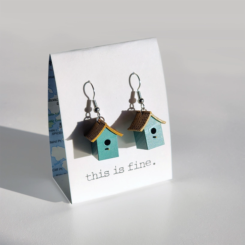 Birdhouse-shaped earrings on a card with 'this is fine' text, against a light gray background
