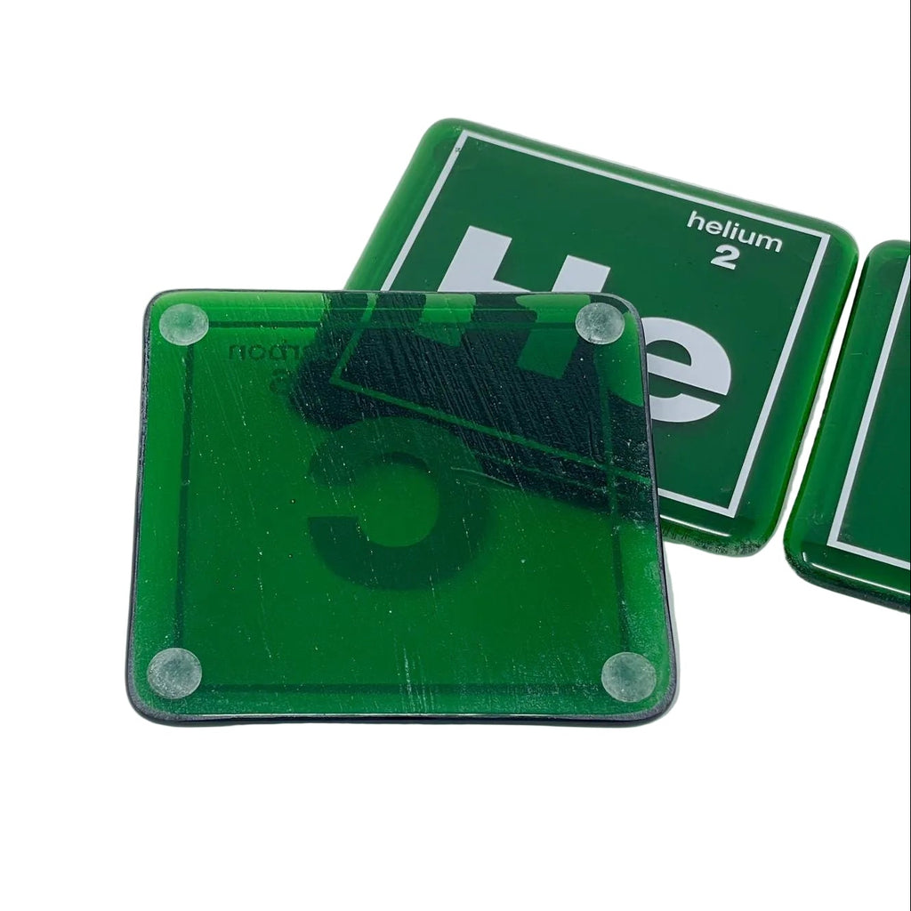 Green periodic table style coasters with rounded corners, shown on a plain white surface. One coaster is placed in front and partially covers another, with a third visible behind it.