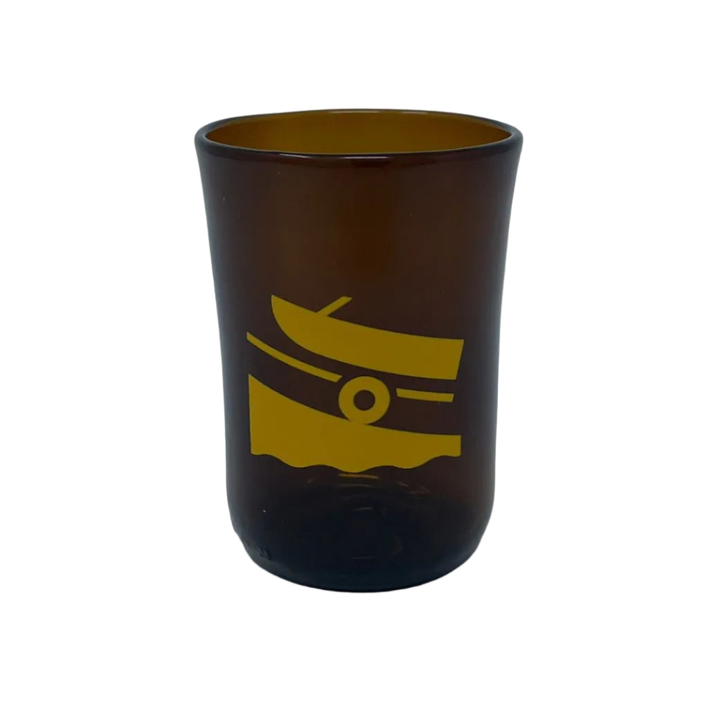 An amber brown glass tumbler with a yellow graphic of a boat on waves, set against a plain light gray background.
