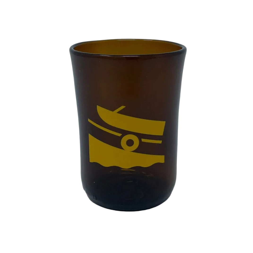 An amber brown glass tumbler with a yellow graphic of a boat on waves, set against a plain light gray background.