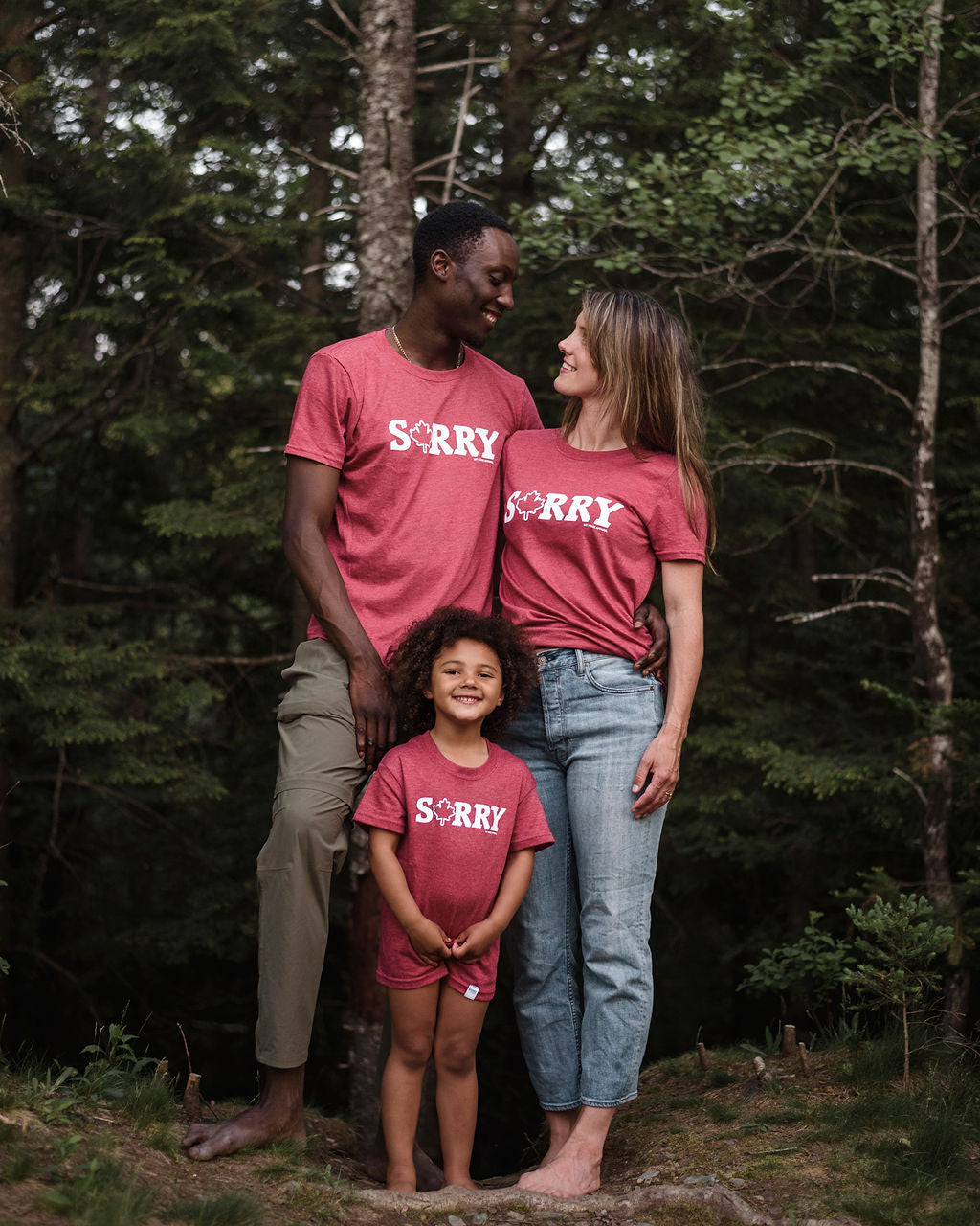 2 adults and one child standing in front of some trees wearing a red t-shirt that reads sorry 