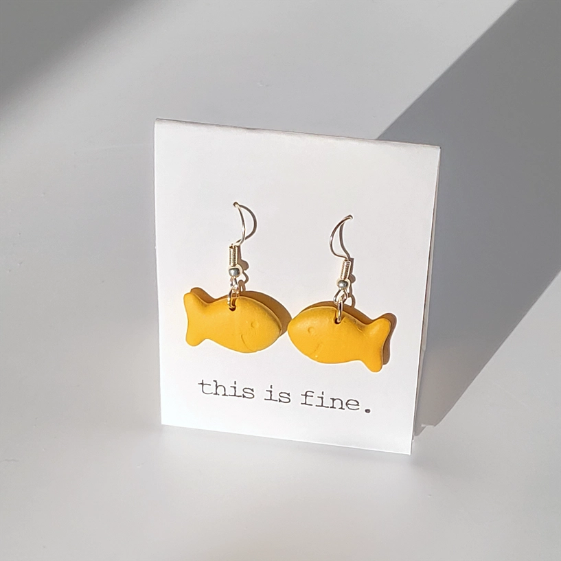 Yellow fish-shaped earrings on a card with 'this is fine' text against a gray background
