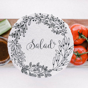 round bowl cover with the word salad written on it & herbal leaves around in front of some tomatoes